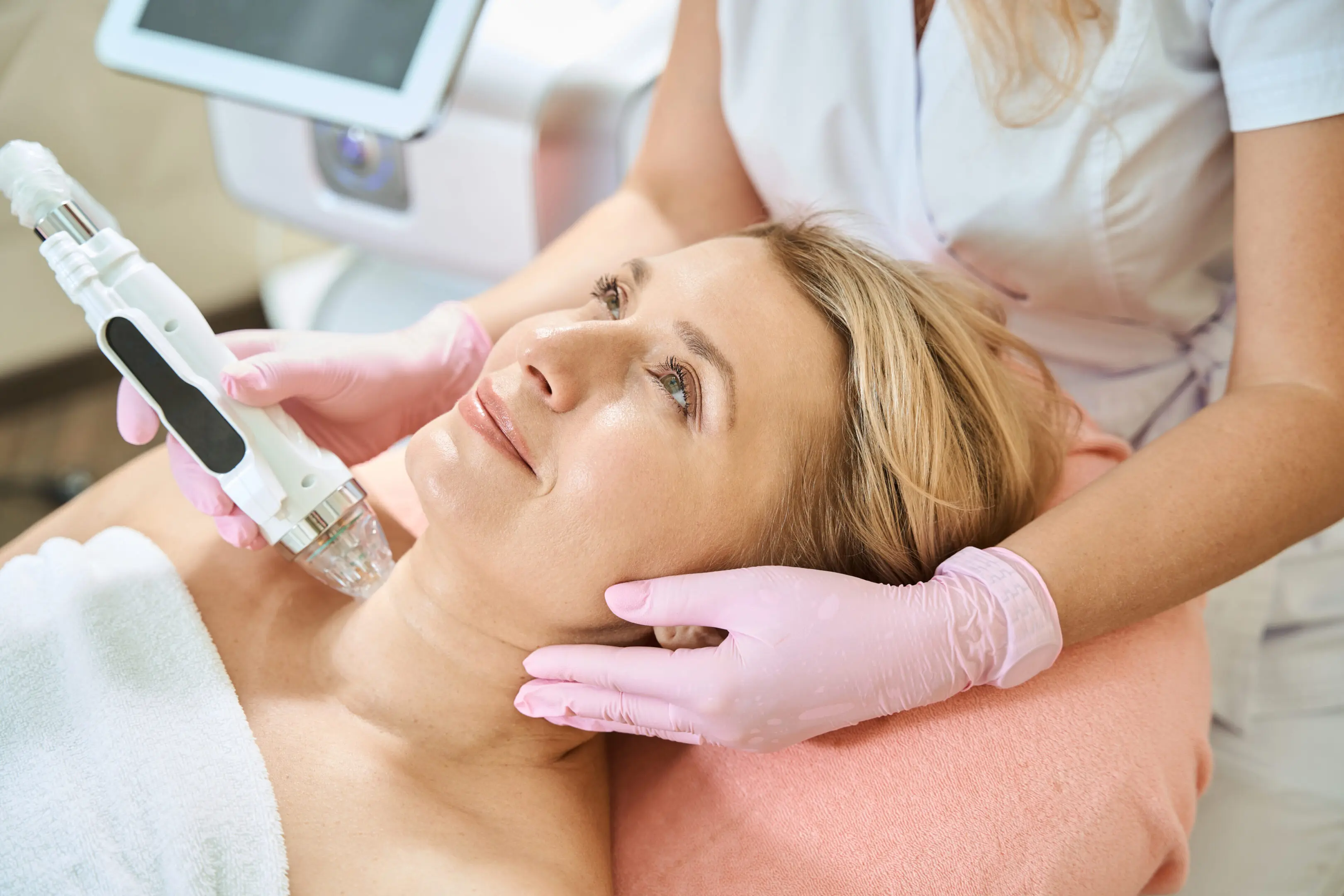 Cropped female cosmetologist doing microneedle rf lifting of neck skin with IPL photorejuvenation device of smiling adult caucasian woman in blurred beauty salon. Body skin care and rejuvenation